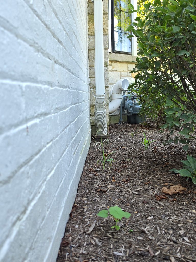 Exterior foundation edge with downspout, utility area, mulch bed, and shrubs on a Chicago-area home