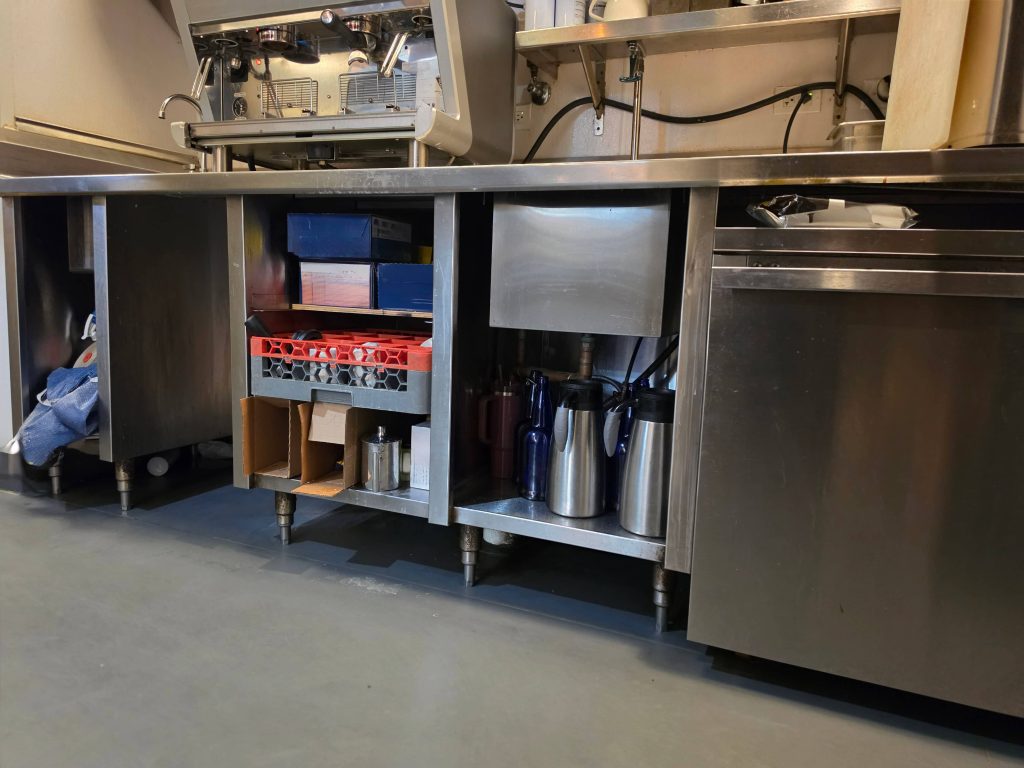 Stainless coffee-service counter with open under-counter shelves and equipment legs above the floor