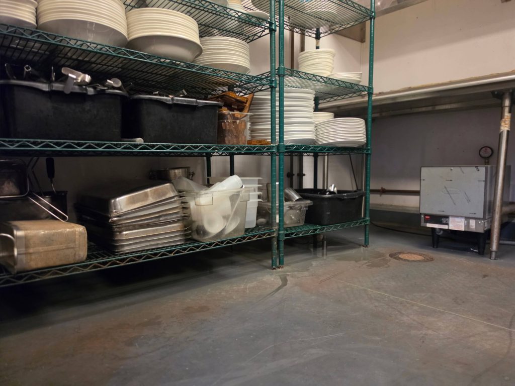 Green wire shelving stacked with plates and bins beside stainless equipment on a commercial kitchen floor